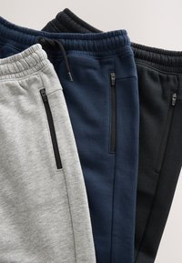 Tre pantaloni della tuta piegati in grigio, blu navy e nero con vita elastica e tasche laterali con cerniera.