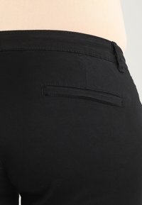 Pantalons en coton noir avec une texture lisse. Comprend une poche arrière discrète et une taille unie. La vue rapprochée met en évidence les détails du matériau et des coutures.