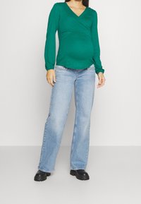 Donna incinta che indossa una maglietta verde a maniche lunghe con scollatura a V e jeans a gamba larga azzurri, in piedi davanti a uno sfondo neutro.