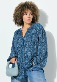 Blusa blu con stampa floreale, con bottoni davanti, maniche lunghe a sbuffo e vestibilità comoda. La modella tiene una piccola borsa in denim.