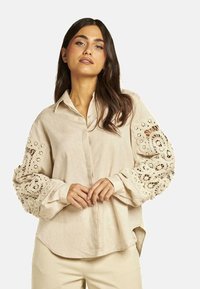 Camisa de lino beige con mangas anchas con encaje, frente abotonado y un corte relajado, con una textura suave y un dobladillo curvado.