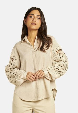 Camisa de lino beige con mangas anchas con encaje, frente abotonado y un corte relajado, con una textura suave y un dobladillo curvado.