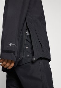 Fekete vízálló dzseki GORE-TEX címkével, cipzárral és patentos záródással, állítható mandzsettákkal és textúrázott szövetfelülettel.