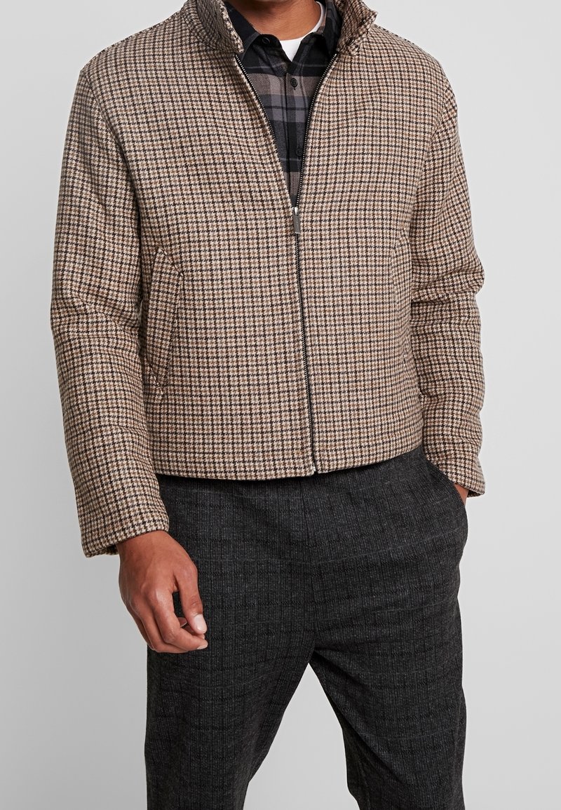 Ruskea ruudullinen puffertakki, jossa on korkea kaulus, etuvetoketju ja kaksi sivutaskua. Pukeutunut mustan ja harmaan ruudullisen paidan päälle.