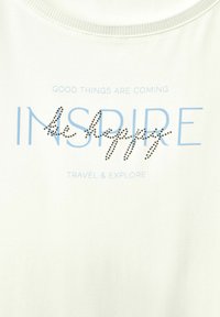 Ljus krämfärgad t-shirt med ribbat halsringning, prydd med blå text "INSPIRE" dekorerad med svarta rhinestones och ytterligare tryckt text.