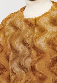 Faux fur-jacka i gyllene-brun med en vågig texturmönster. Har en hög krage och mjukt, plysch material. Närbild av tygdetalj.