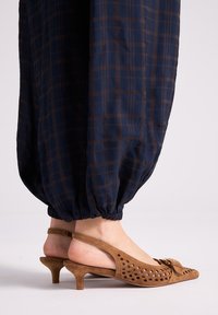 Pies con zapatos de tacón bajo tipo kitten slingback de ante marrón con patrón recortado y lazo, combinados con pantalones de puño elástico a cuadros azul oscuro.