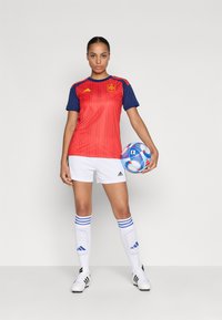 Woman in red and blue soccer jersey, white shorts, and socks holding a blue and white soccer ball, standing against a plain background.