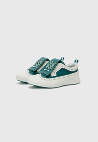 Baskets avec un design blanc et teal, présentant un dessus texturé, des perforations décoratives et de larges sangles teal avec un logo.