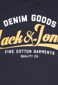 Tmavě modré bavlněné svetry s bílým a žlutým textem "DENIM GOODS Jack & Jones" a jemnými bavlněnými oděvy níže.