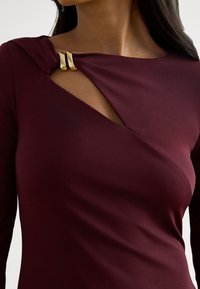 Vijolično rdeča obleka z dolgimi rokavi, zasukanim izrezom okoli vratu in zlato opremo na ramenih ter gladko, sijočo teksturo.