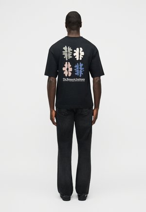 Homme debout de dos portant un t-shirt noir avec quatre motifs de cerveau abstraits en gris, rose, bleu et blanc, accompagnés d'un texte en dessous.