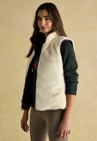Svetle krémová vesta s přední částí z umělé kožešiny, tmavým lemem a bezrukávovým designem. Přes tmavý pletený svetr a vzorované kalhoty.