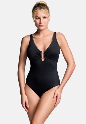 Frau mit blondem Haar zu einem Dutt frisiert, die einen schwarzen Badeanzug mit tiefem V-Ausschnitt und einem Metallring vorne trägt, steht vor weißem Hintergrund.