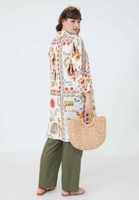 Long, loose white tunic with colorful prints, including flowers and phrases, paired with olive green wide-leg pants. Woven round handbag in hand.