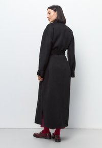 Palton lung negru cu talie evidențiată prin curea, mâneci largi, slituri laterale și o textură netedă. Purtați peste o rochie burgundi și asortați cu pantofi loafers.