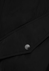 Prenda exterior negra con acabado mate elegante, ángulos rectos y un detalle metálico de botón a presión con el grabado "Polar Bear". Textura suave, diseño minimalista.