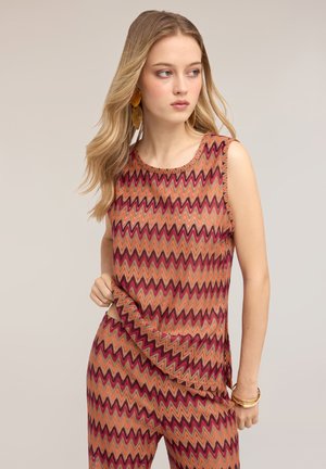 Jonge vrouw met lang blond haar draagt een mouwloze top met zigzagpatroon en bijpassende broek in warme tinten, poserend met hand in de taille.