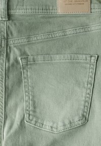 Hellgrüner Denim-Stoff mit einer hinteren Tasche, die mit horizontalen Nähten versehen ist, und einem darüber angebrachten Lederetikett, das den Markennamen und den Slogan zeigt.