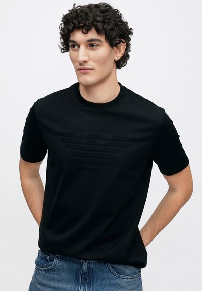 Μαύρο βαμβακερό μπλουζάκι με ανάγλυφο λογότυπο αετού σε τονικό χρώμα, στρογγυλή λαιμόκοψη, κοντά μανίκια και ίσια γραμμή.