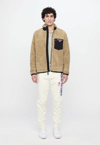 Man standing wearing beige fleece jacket with black chest pocket, white sweatpants with blue "Polo Sport" text, and white sneakers.