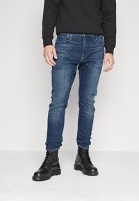 Uske slim-fit traperice tamnoplavog tona imaju pet džepova i zatvarač. Uparen s crnim vojničkim čizmama za kontrast. Materijal je mješavina pamuka.