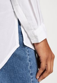 Gros plan sur une main reposant près de la poche d'un jean en denim bleu, portant une chemise blanche à manches longues avec un poignet boutonné et une fente latérale.