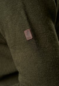 Pull en maille vert olive avec un tissu texturé, orné d'un petit patch en cuir marron affichant un logo cousu sur la manche.