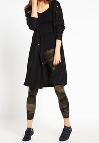 En kvinna står självsäkert, klädd i en svart linne, en lång svart jacka och kamouflageleggings, tillsammans med marinblå skor.