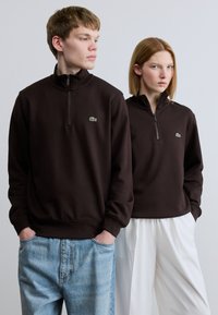 Καφέ φούτερ με φερμουάρ από απαλό ύφασμα, με μικρό πράσινο λογότυπο, φορεμένα με ανοιχτόχρωμο μπλε τζιν και λευκό παντελόνι με φαρδιά μπατζάκια.