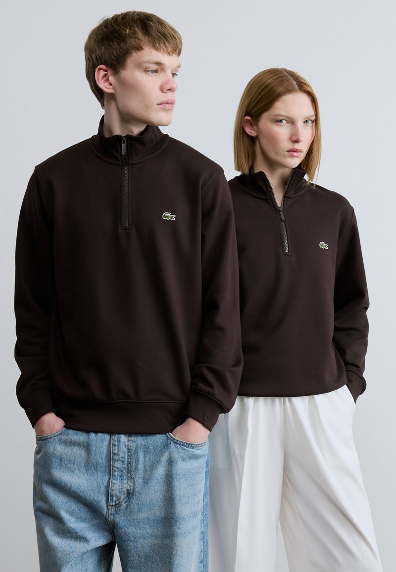 Καφέ φούτερ με φερμουάρ από απαλό ύφασμα, με μικρό πράσινο λογότυπο, φορεμένα με ανοιχτόχρωμο μπλε τζιν και λευκό παντελόνι με φαρδιά μπατζάκια.