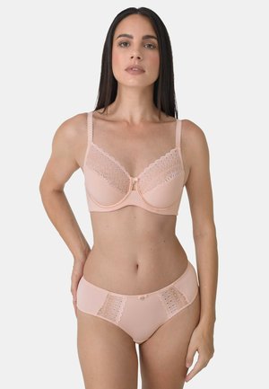 Frau, die passende hellrosa Unterwäsche mit Spitzenbesatz trägt, bestehend aus BH und Slip, steht mit einer Hand in der Hüfte vor einem schlichten Hintergrund.