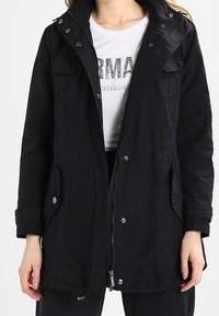 Parka negra con cremallera frontal, bolsillos abotonados y un cordón ajustable en la cintura. Presenta una capucha y una textura de tela suave.