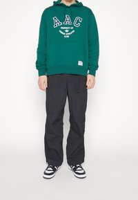 Grön hoodie med "AAC"-logotyp, framficka och justerbar dragsko. Svarta, löst sittande byxor och svarta och vita sneakers fulländar outfitten.