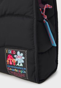 Svart polstret veske med fargerik brodert lapp som viser sopp og en blomst, samt et blått merke med teksten "Coachtopia." Teksten inneholder lekne setninger.