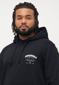 Man with beard and dreadlocks wearing a black hoodie with a small white "Originals International" logo and text.