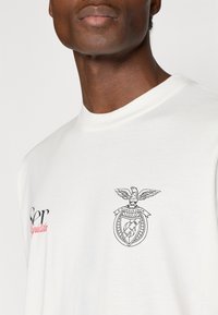 Biała bawełniana koszulka z czarną grafiką orła i tarczy na klatce piersiowej, z napisem "Ser" w czerwieni powyżej. Luźny krój i okrągły dekolt.