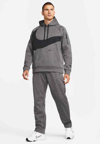 Homme portant un sweat à capuche Nike gris et un pantalon de survêtement assorti avec un grand swoosh noir, debout avec des baskets Nike blanches sur fond blanc.