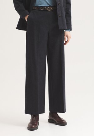 Personne portant un pantalon large foncé, une ceinture cloutée, un haut bleu, une veste et des bottines marron foncé sur un fond clair.
