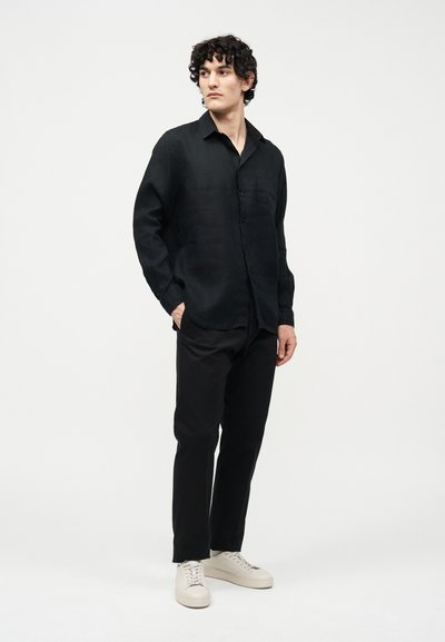 Άνδρας με σγουρά μαλλιά, φορώντας μαύρο πουκάμισο με κουμπιά, μαύρο παντελόνι και λευκά αθλητικά παπούτσια, στέκεται με το ένα χέρι στην τσέπη του.