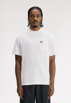 Homme aux cheveux tressés portant une chemise blanche à manches courtes avec une poche poitrine et un logo brodé noir, assortie à un pantalon foncé.