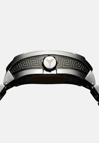 Silver metal watch with a textured black band and engraved Greek key pattern. Features a round crown with a logo at the center.