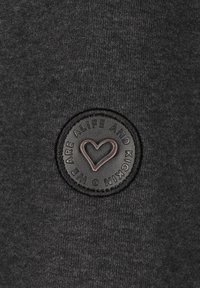 La tela del sudadera gris oscuro presenta un parche circular negro texturizado con letras en relieve y un diseño de corazón en el centro.