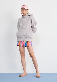 Šedá oversized mikina s přední kapsou, růžová kšiltovka a barevné šortky s proužky a tropickým vzorem. Béžové sandály.