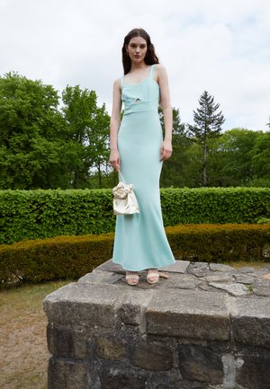 Vestido justo verde menta com um recorte, alças finas, combinado com uma bolsa de cetim creme e sandálias nudé, de pé sobre uma plataforma de pedra.