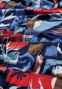 Tessuto di cotone con un motivo astratto multicolore che presenta toni di blu navy, rosso, blu e marrone, mostrando forme che ricordano foglie e blocchi.