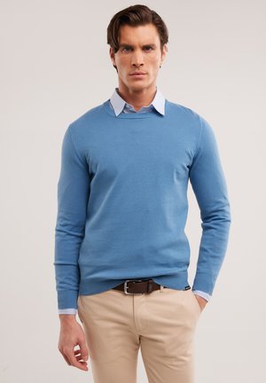 Maglione lavorato a maglia azzurro con scollatura rotonda, polsini a coste e una texture morbida, indossato sopra una camicia azzurra con colletto e pantaloni beige.