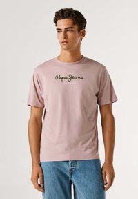 Svetlo roza bombažna majica s kratkimi rokavi, ki ima zeleni logo "Pepe Jeans" in besedilo "LONDON" centrirano spodaj od logotipa.