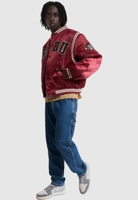 Röd satäng varsityjacka med svarta bokstäver och randiga muddar, tillsammans med ljusblå denimjeans och grå sneakers.