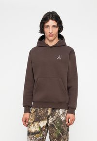 Joven vestido con una sudadera marrón con un pequeño logo blanco en el pecho y pantalones de patrón camuflaje, de pie contra un fondo blanco liso.